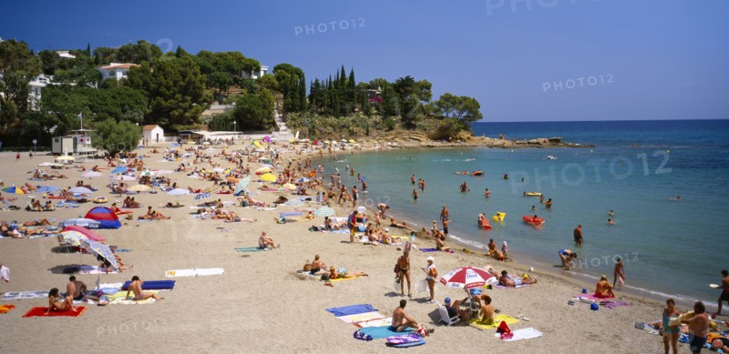 SPAIN, Catalonia, Llanca, "Curved, sandy beach with people sunbathing and in the sea.  White painted houses amongst trees on hillside beyond."
