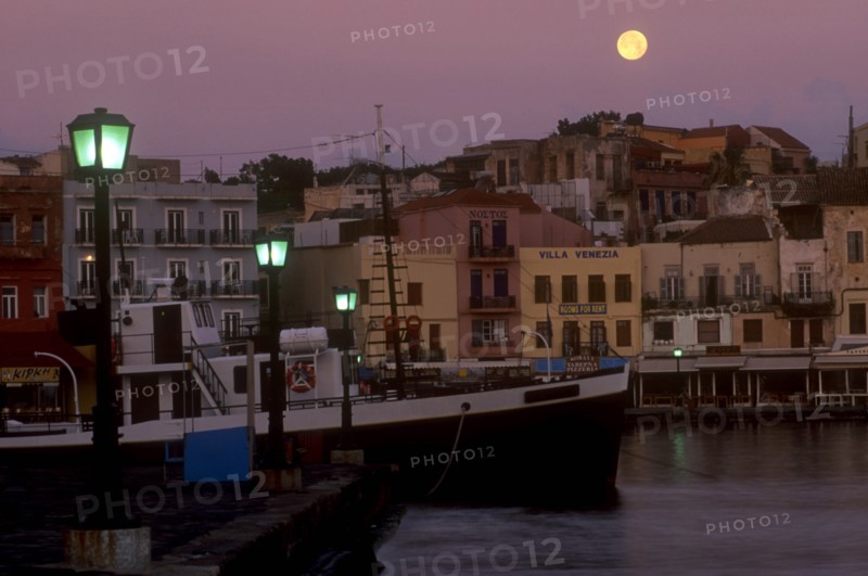 GREECE, Crete, Hania, Venetian Port.  Moored boat and quayside buildings with moon in pale purple dusk sky above and illuminated street lamps.