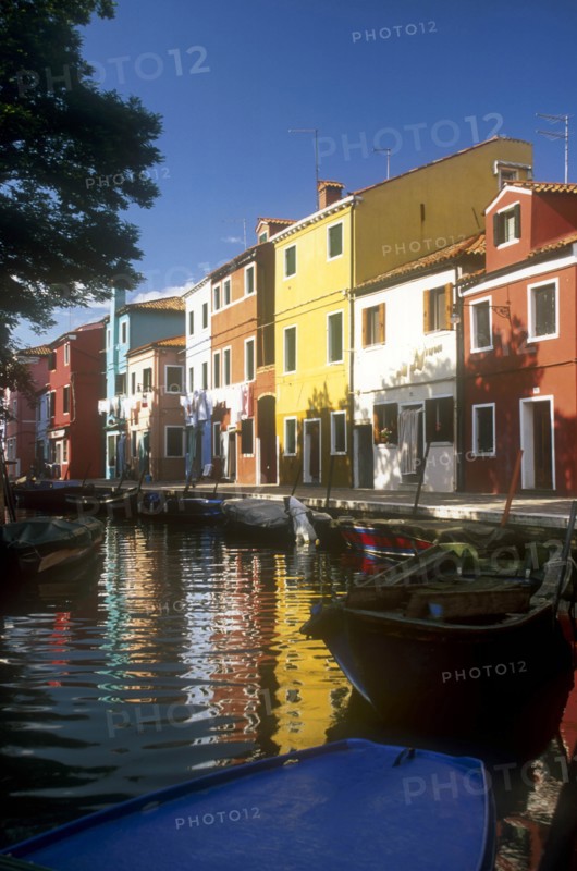 ITALY, Veneto, Venice, Burano Island.  Boats moored at canal-side overlooked by row  of brightly painted buildings.