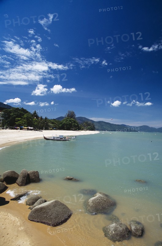 MALAYSIA, Penang, Batu Ferringhi, View along shore of quiet sandy beach with moored boat in middle distance and smooth rocks in shallow water in the foreground.