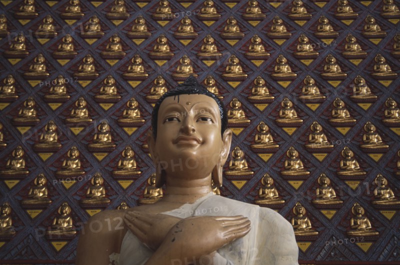 MALAYSIA, Penang, Georgetown, Wat Chayamangkalaram.  Detail of Buddha figure with hands crossed over chest.  Wall behind covered with small raised seated Buddha figures painted gold.