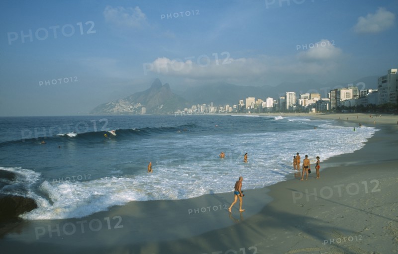BRAZIL, Rio State, Rio de Janeiro, Ipanema Beach with bathers standing at the waters edge in early morning light with misty mountains in the distance.