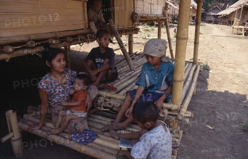 THAILAND, North, Mae Sai , Karen refugee mother feeding child with four other children sitting in the shade