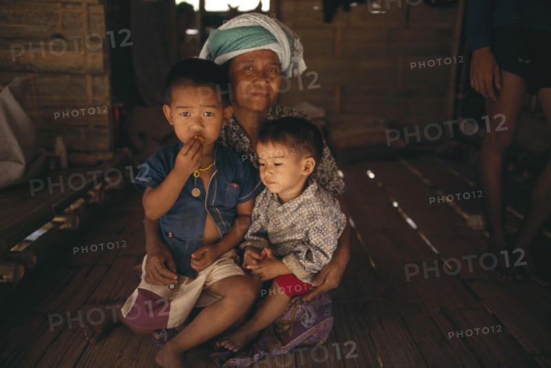 THAILAND, North, Mae Sai Area, Karen Refugee mother sitting with her two children on the floor of a hut