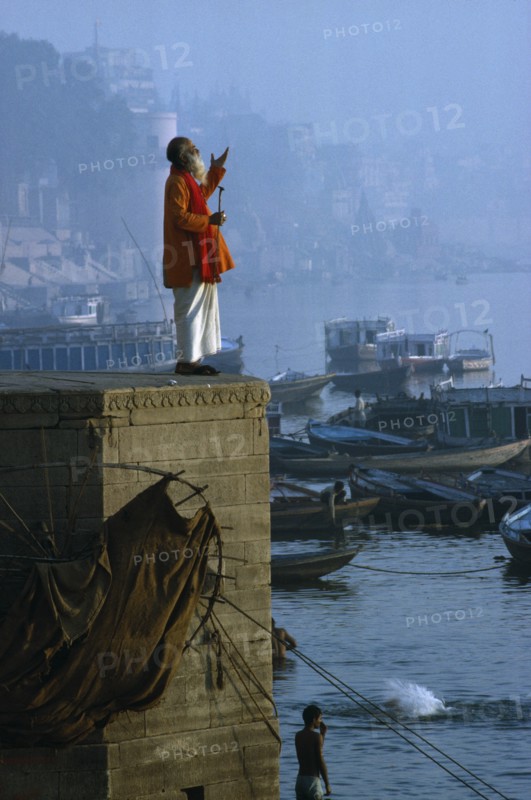 INDIA, Uttar Pradesh, Varanasi, "Holy man praying on stone wall above River Ganges,boats,town,mist "