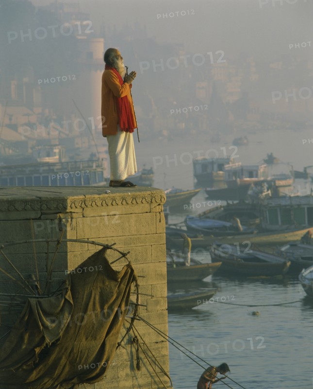 INDIA, Uttar Pradesh, Varanasi, Holy man praying on stone wall above River Ganges with misty town and boats in background