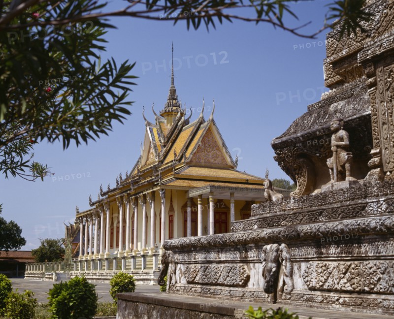 CAMBODIA, Phnom Penh, Silver Pagoda.  Exterior with ornate golden roof with spire and stone monument in foreground.