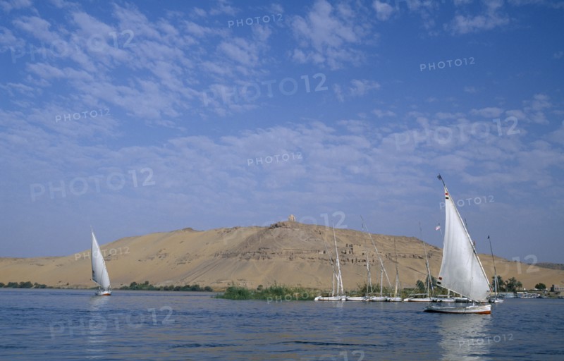 EGYPT, Upper Egypt, Aswan, Qubbet el-Hawa and The Tombs of The Nobles with two feluccas sailing past on the river Nile