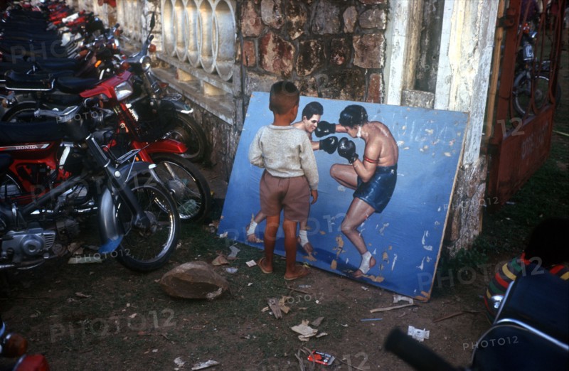 CAMBODIA, Battambang, Small boy looking at painting of boxers propped against the wall in a motorcycle yard.