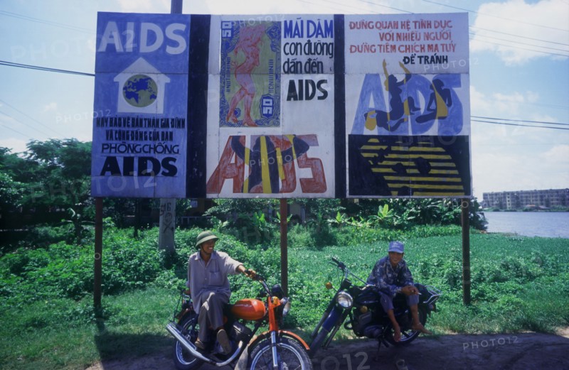 VIETNAM, Hai Duong, Two young men sitting on their motorcycles under a billboard of aids awareness posters.