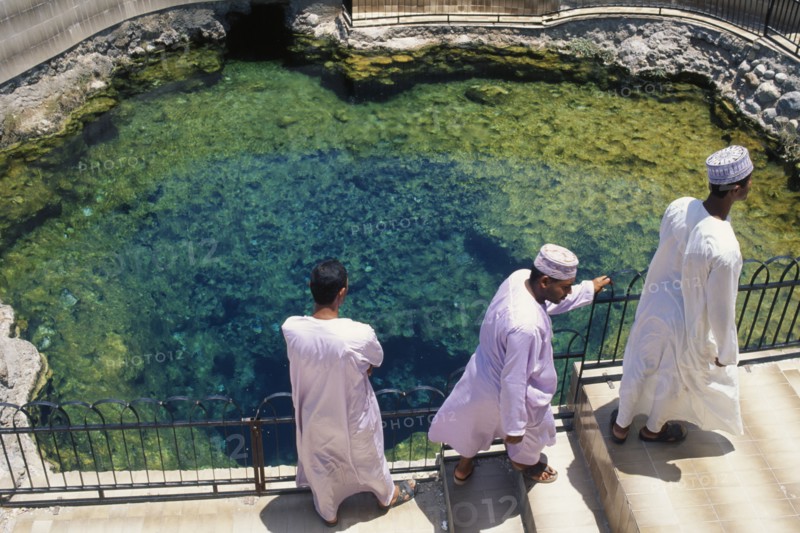OMAN, Al Rustaq, "Three men in traditional white dishdashas and brimless, circular hats standing beside hot springs."