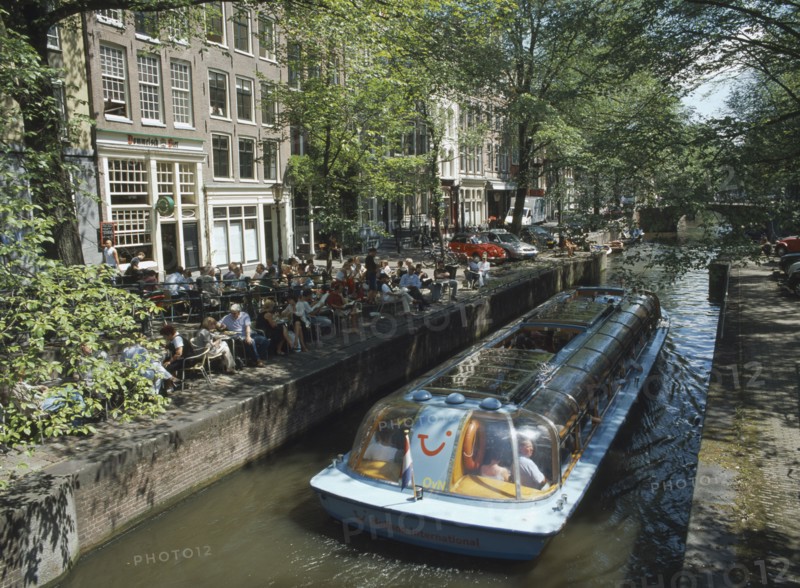 HOLLAND, Noord, Amsterdam, Glass roofed tourist boat passing canal side cafe on the Lelegracht canal