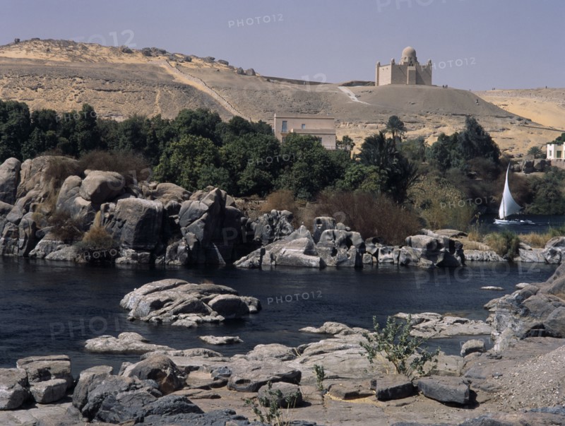 EGYPT, Nile Valley, Aswan, Feluccas on the Nile. View of Felucca sailing among the boulders off  Elephantine Island with the Mausoleum of the Aga Khan in the background.