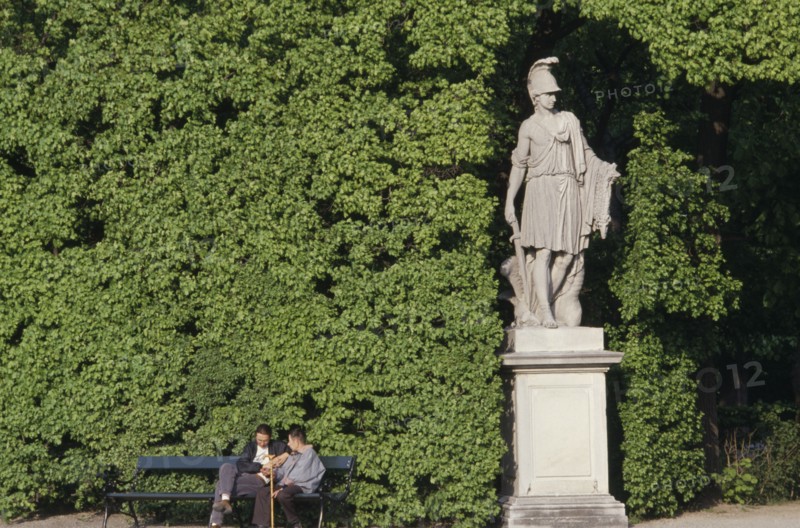 AUSTRIA, Vienna, Schonbrunn Gardens.  Classical statue against hedge with two visitors sitting on bench at one side.