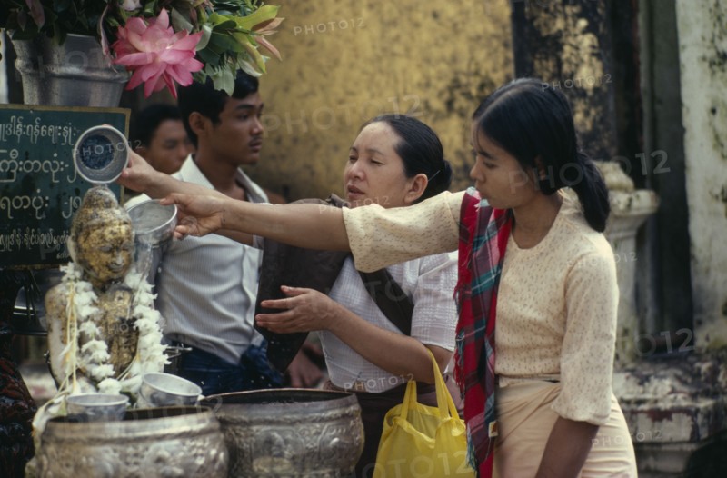 MYANMAR, People, Young women making offerings at Buddhist shrine with small seated Buddha figure garlanded with flowers.