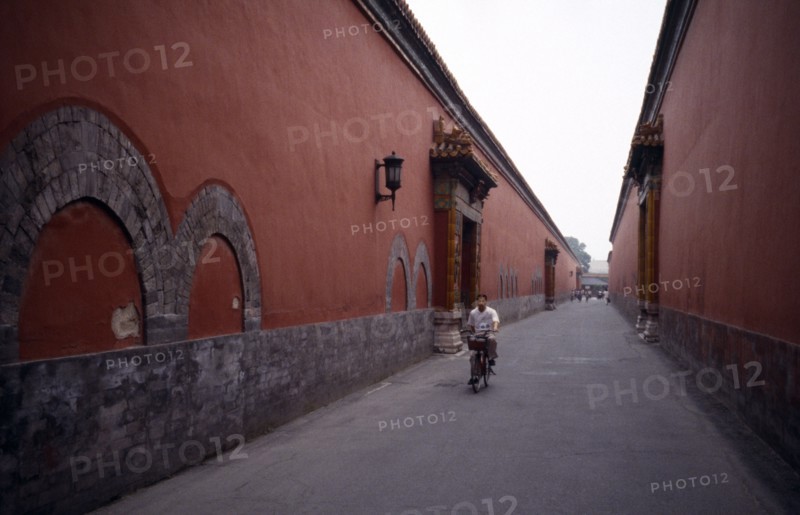 CHINA, Beijing, The Forbidden City.  Cyclist on narrow street between red painted walls.