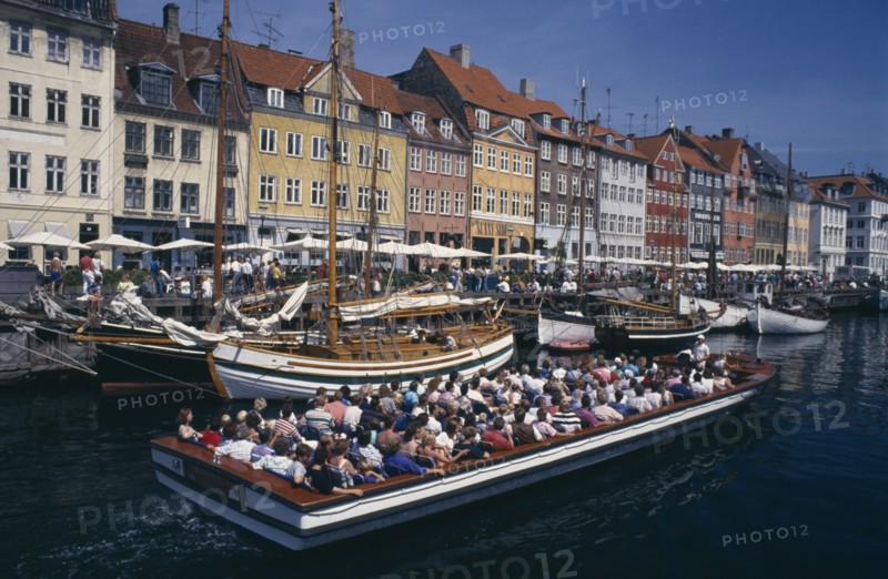 DENMARK, Zealand, Copenhagen, Nyhavn Canal. Cruise boat passing sailing boats moored on the quayside beside brightly painted traditional houses