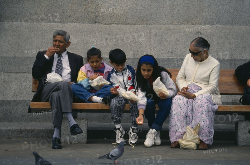 ENGLAND, London, Trafalgar Square, Asian family sitting together on a bench eating with girl leaning forward to feed a pigeon