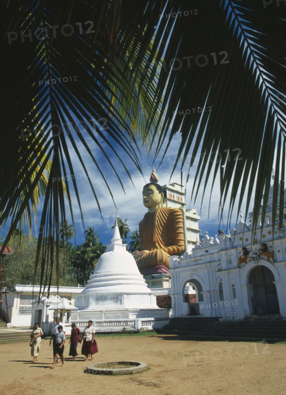 SRI LANKA, Wewurukannala Vihara, Site of giant seated Buddha constructed in 1970 with visiting family in courtyard in foreground.  Part framed by palm fronds.