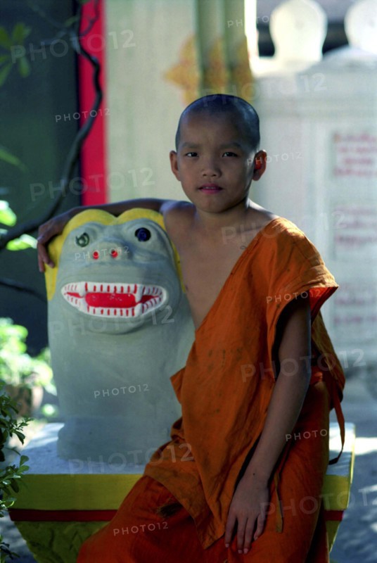 LAOS, Savannakhet, Wat Sainyamungkhun. Young monk sitting beside colourful statue at the Temple