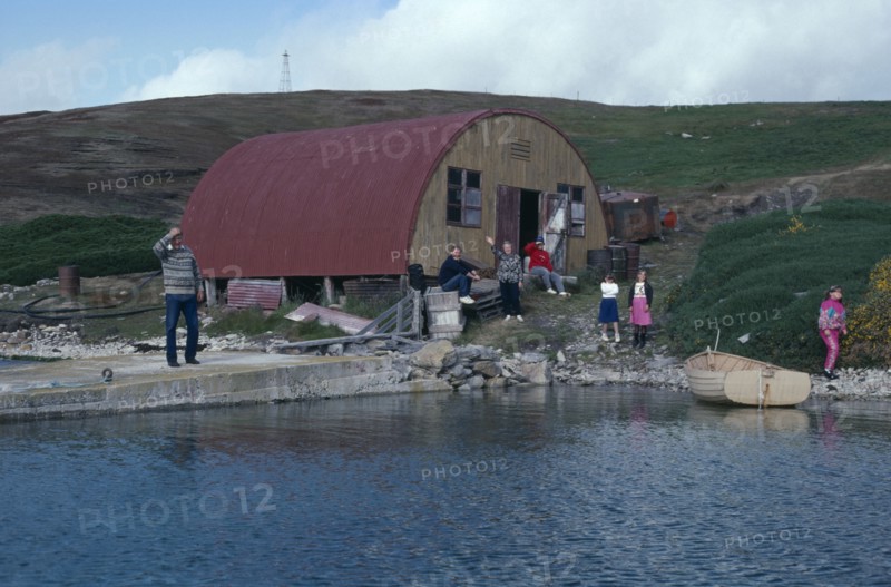 FALKLAND ISLANDS, West Point Island, View across water towards loading jetty with a family near building
