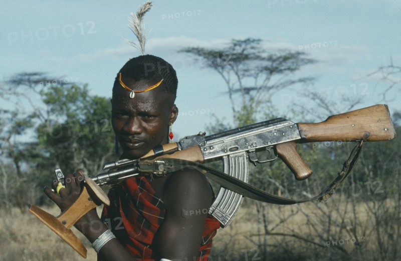 UGANDA,   , Karamoja, "Portrait of Karamojong warrior with ostrich feather in hair holding gun and stool, always carried and used as both seat and head rest. "