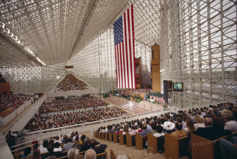 USA, California, Los Angeles, Crystal Cathedral Memorial Service. Congregation in pews under glass structure beneath flag of Stars And Stripes and a large TV