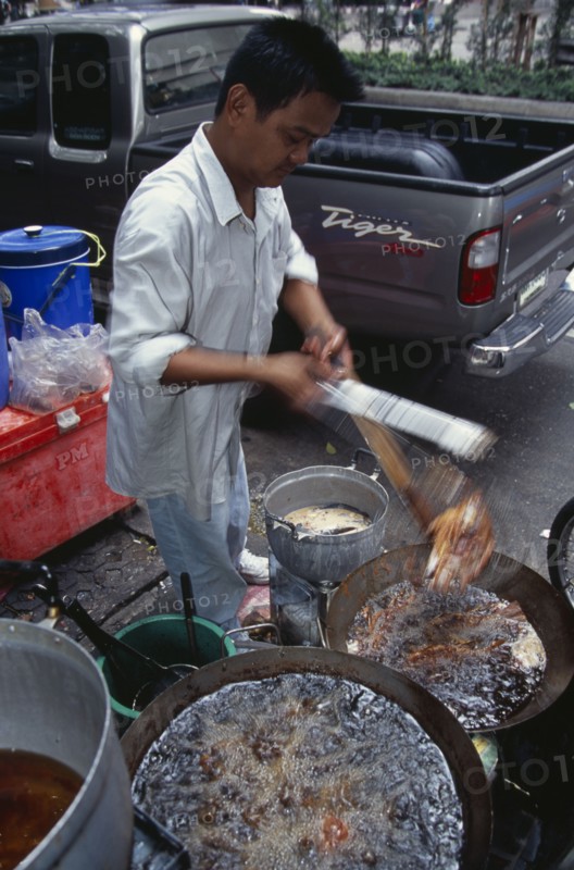 THAILAND, South, Bangkok, Thanon Silom male vendor at his stall on the pavement deep frying fish in woks