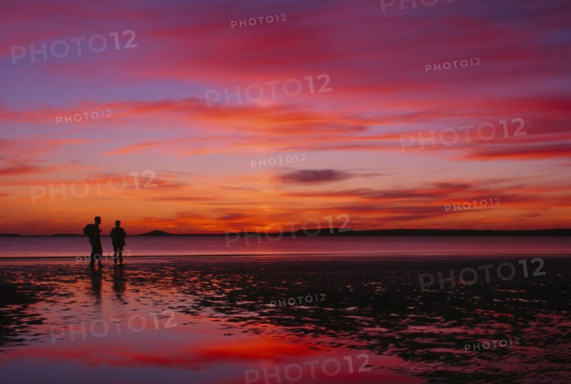 IRELAND, Sligo, Rosses Point, Two figures silhouetted on the beach watching dramatic deep pink and orange sunset reflected on wet sand.