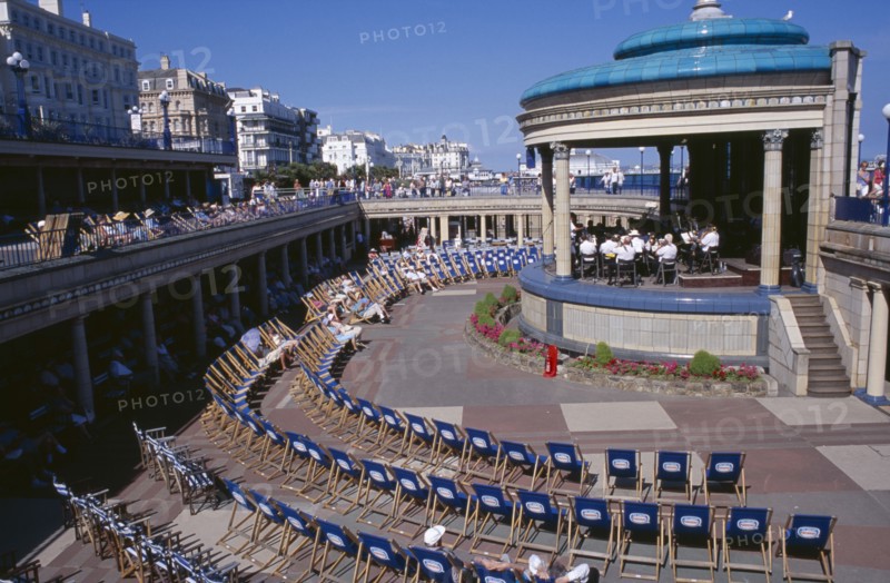 20059185 ENGLAND East Sussex Eastbourne View towards the Promenade Band Stand with a brass band performing on a sunny day and spectators watching from deckchairs layed out in a semi-circle.