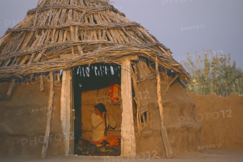 INDIA, Rajasthan, Thar Desert, Bhikodai.  Early morning light on thatched mud hut with woman plaiting her hair framed in doorway.