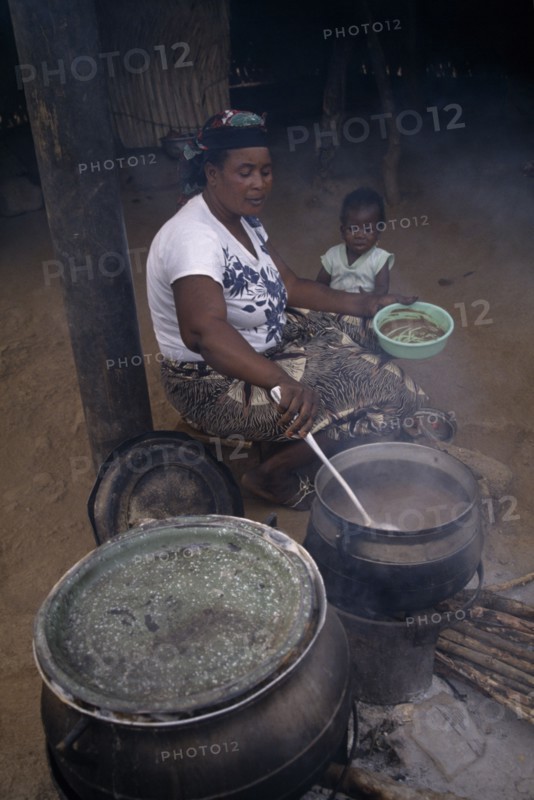 GHANA, Cherepani, Woman cooking over wood burning open topped stove and passing helping of food to small girl sitting beside her.
