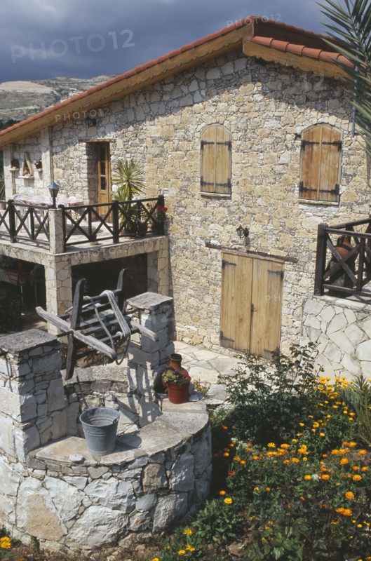 CYPRUS, Lineia, Stone house with overhanging tiled roof and wooden window shutters and door with well in courtyard in the foreground.