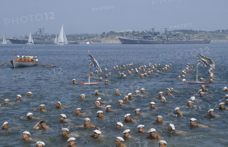 RUSSIA, Sevastopol, Navy Day. Sailors swimming in the sea in formation with battleships behind.