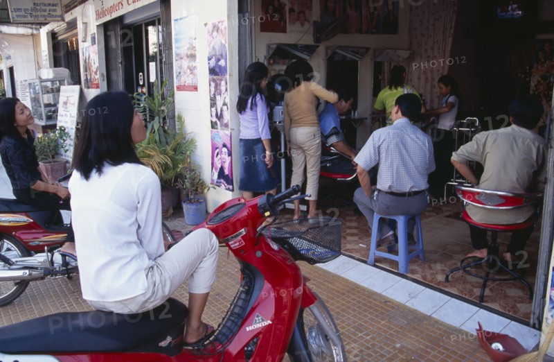 CAMBODIA, Siem Reap, Busy hairderssers shop with two women sitting on mopeds watching a television in the back of the shop