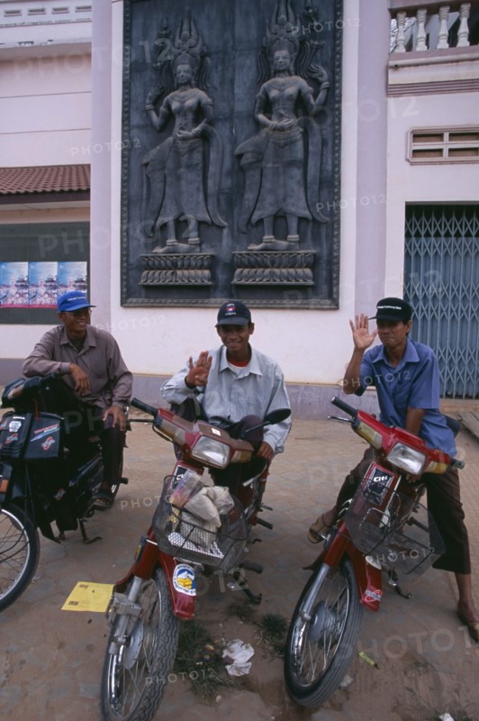 CAMBODIA, Siem Reap, Three motorbike taxi drivers on their mopeds beneath a large wall plaque depicting Asparas