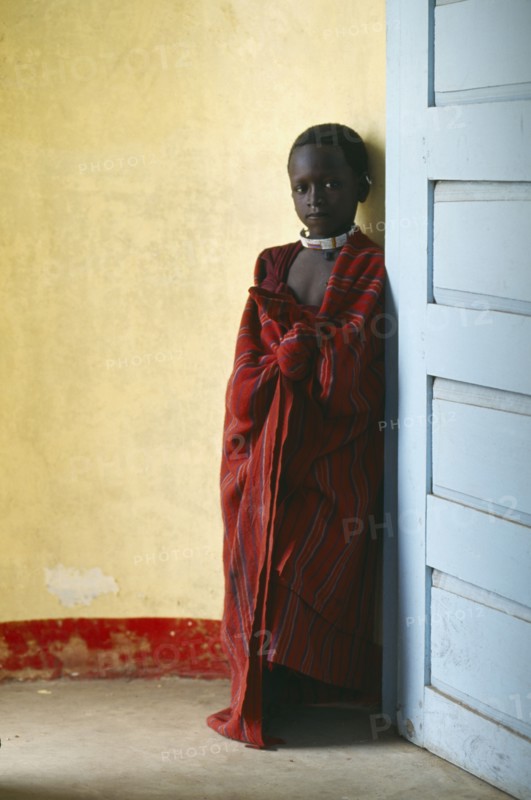 TANZANIA, Ngorongoro, Masai child standing between yellow wall and blue door.  The Masai have been expelled from the Ngorongoro Crater area to make way for tourists and wildlife.
