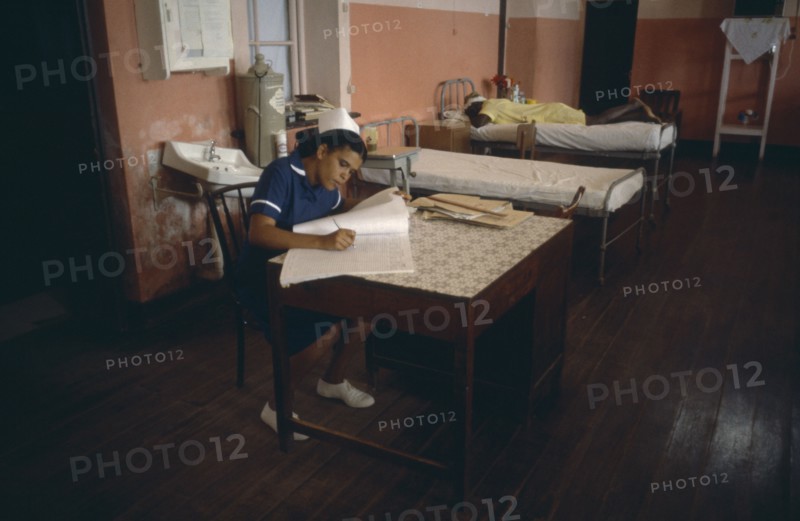WEST INDIES, Grenada, Health, Hospital interior with nurse writing notes and patient lying on bed behind.