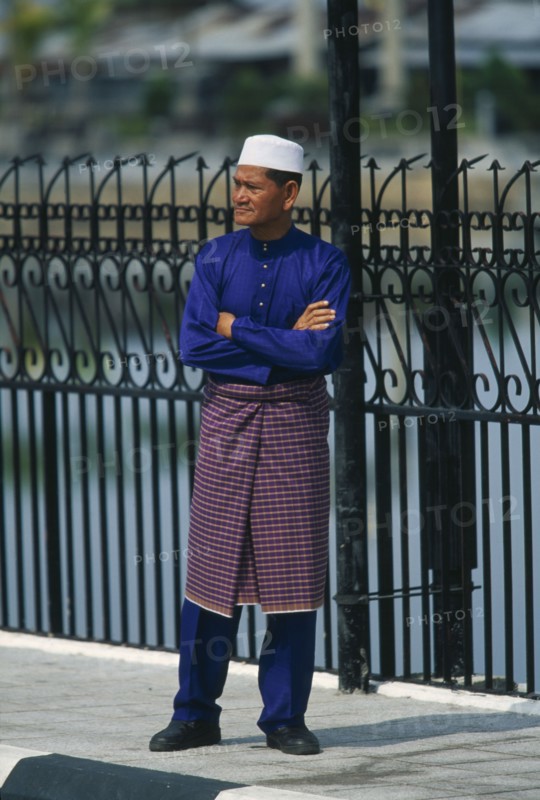 BRUNEI, People, Men, Man standing on pavement with arms crossed next to railings dressed up to visit the Omar Ali Saifuddin Mosque in Bandar Seri Begawan.