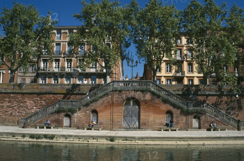 FRANCE, Midi Pyrenees, Haute Garonne, Toulouse.  Red brick steps leading down to the River Garonne from street level with facades of buildings partly seen through trees behind.  People sitting in the sun.
