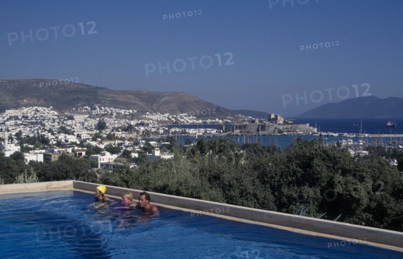 TURKEY, Aegean Region, Mugla, Bodrum.  People in swimming pool of Antik Theatre Hotel in foreground with view over resort to fifteenth century castle of St Peter beyond.