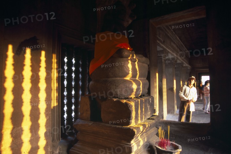 CAMBODIA, Siem Reap Province, Angkor Wat, Tourist looking into interior of shrine in upper or third level with Buddha figure seated below a naga and incense offerings buring at base.