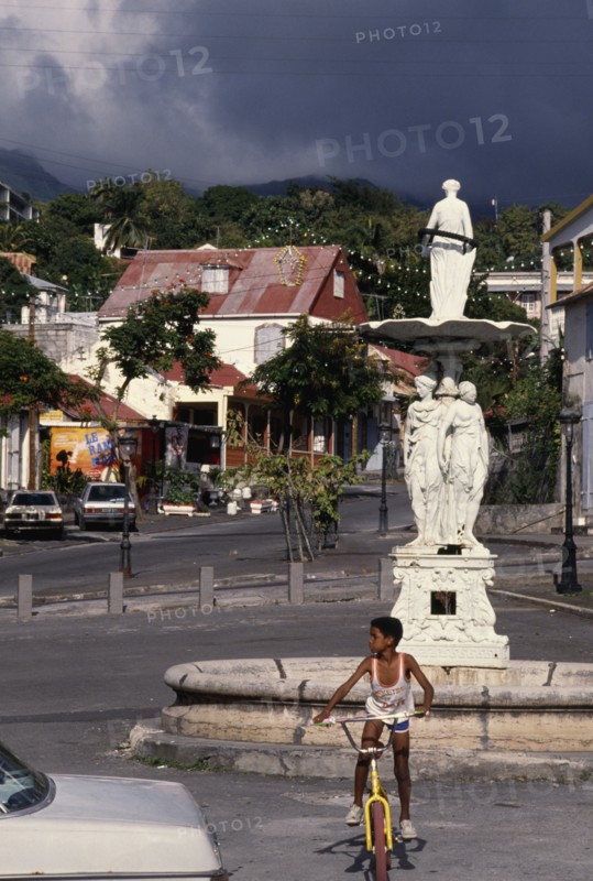 WEST INDIES, Guadeloupe, Basseterre, Large white statue in middle of street surrounded by houses and a boy on a bike in the foreground.