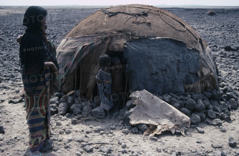 ETHIOPIA, Danakil Depression, Afar woman carrying child standing outside hut or ari with two young children in the doorway n barren arid landscape.