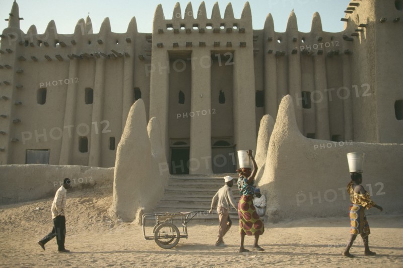 MALI, Djenne, Exterior of mud brick mosque built in traditional style with woman walking past carrying buckets on their heads.