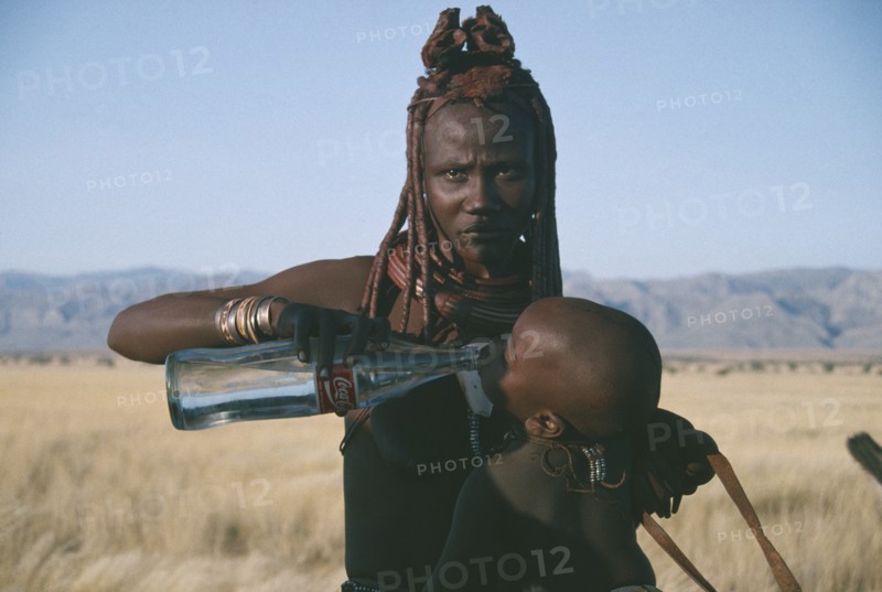 NAMIBIA, Marienfluss, Himba woman giving child a drink of water from Coca Cola bottle.  The Himba traditional way of life is being threatened by uncontrolled tourism.