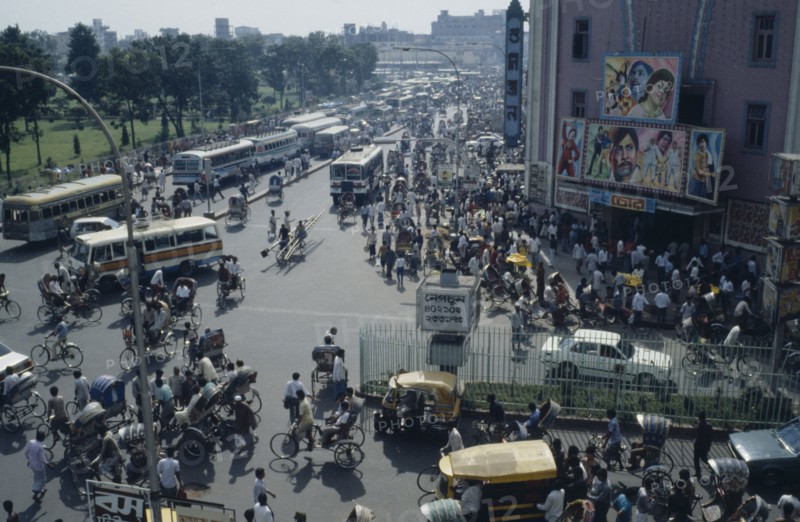 BANGLADESH, Dhaka, Busy street scene with crowds of pedestrians trishaws and buses.  Cinema with billboard advertising films on right.