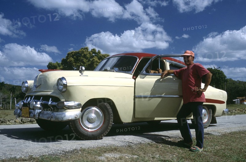 CUBA, Cienfuegos, Man standing by the open door of a classic style American car which has been fitted with tractor tyres due to a tyre shortage