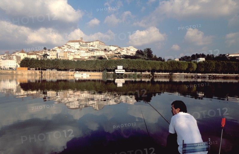 PORTUGAL, Coimbra, Man fishing on the bank of the River Mondego with the University crowning Alcacova Hill opposite