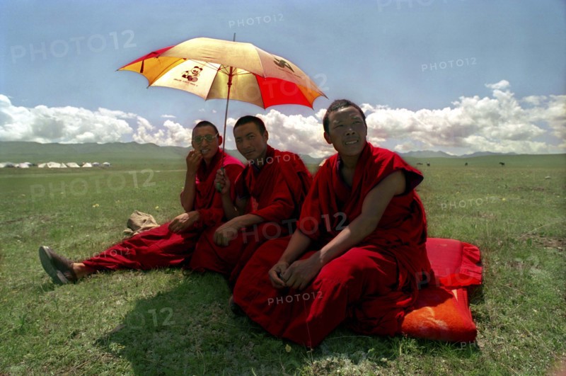 CHINA, Gansu, Xiahe, Three monks with umbrella sitting in green open landscape watching festival